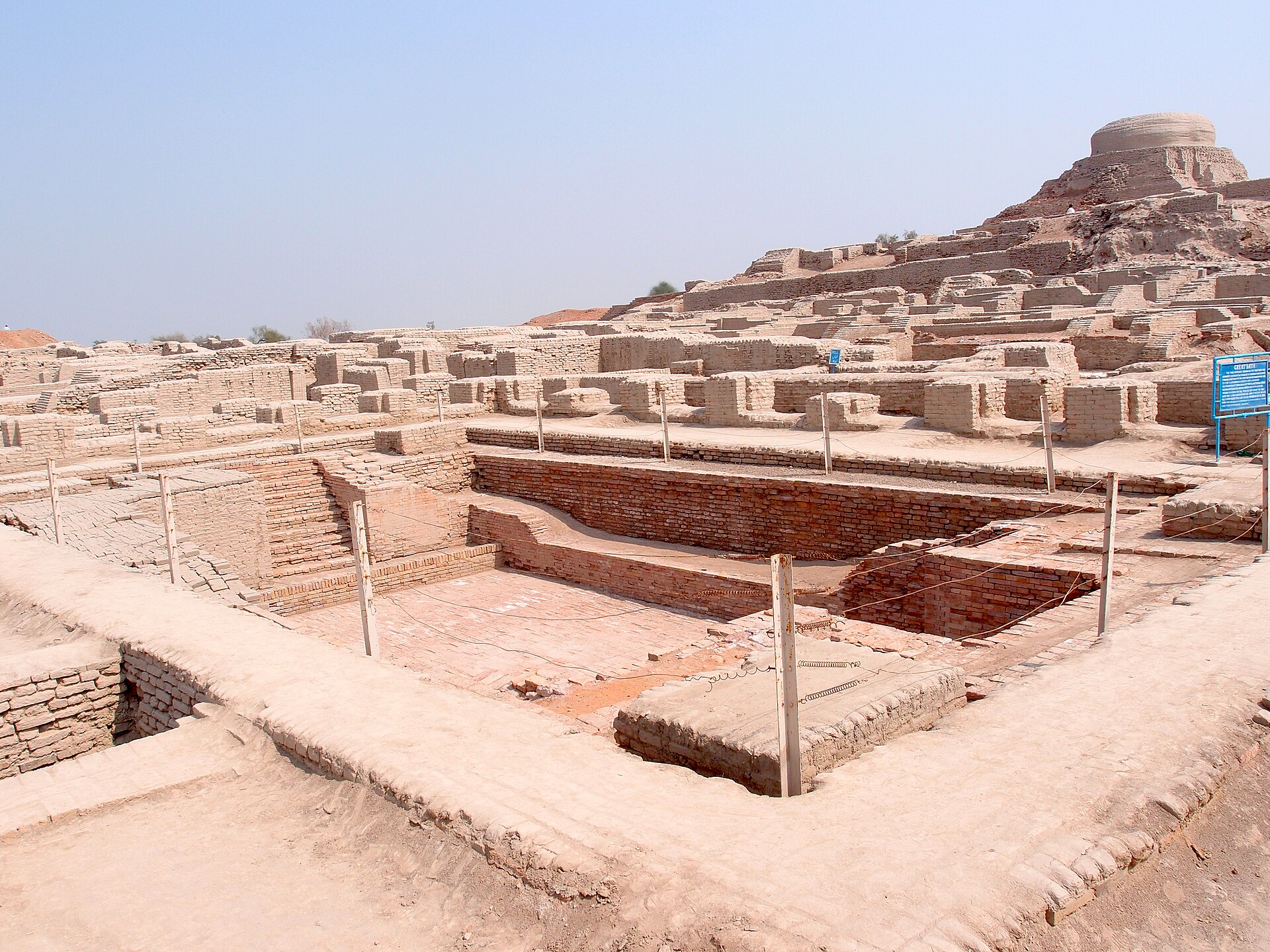 The excavated brick ruins of Mohenjo-daro under a hazy sky, with a later Buddhist stupa rising on a mound in the background