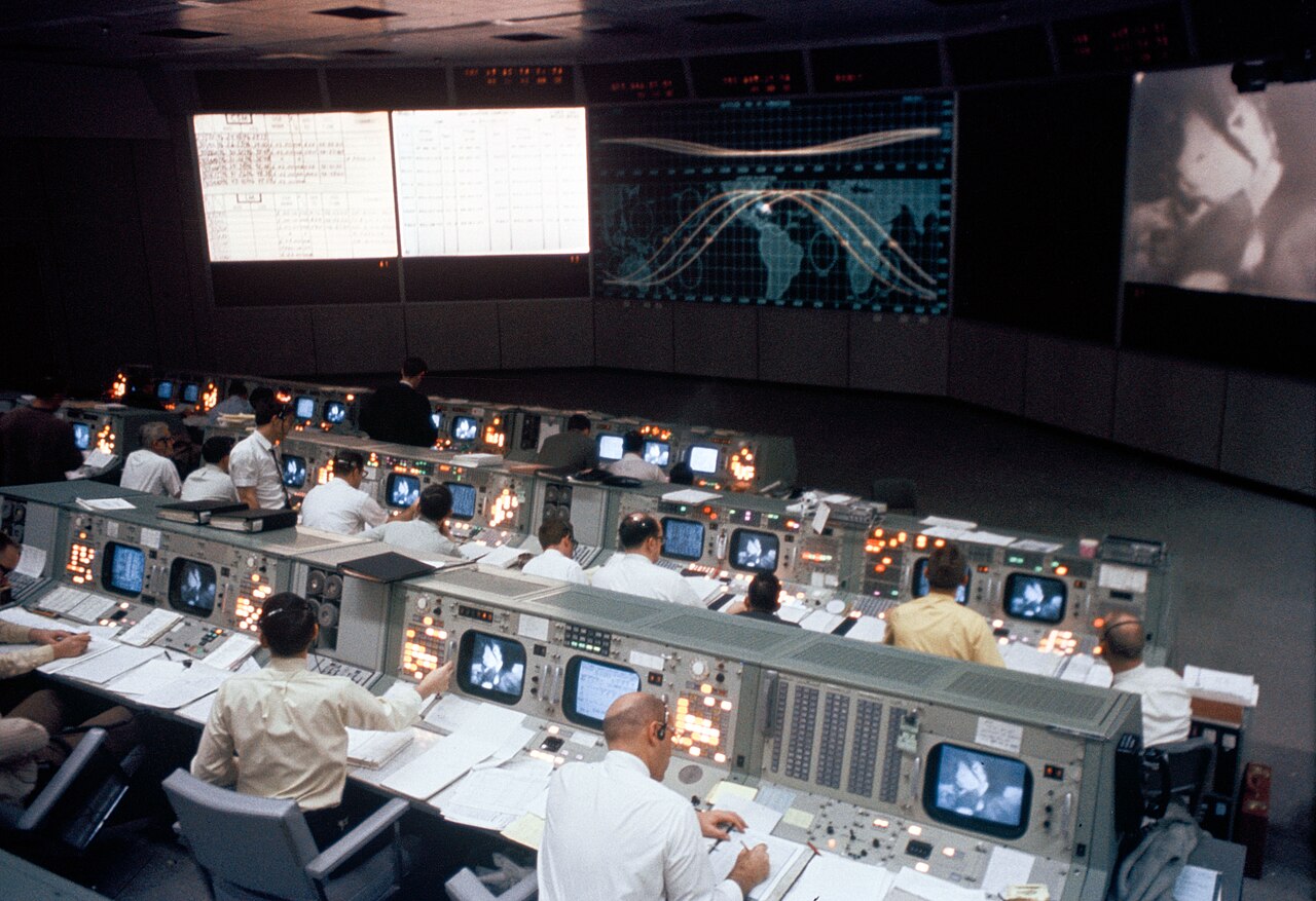 Rows of operators seated at consoles in a large mission control room, with display screens at the front
