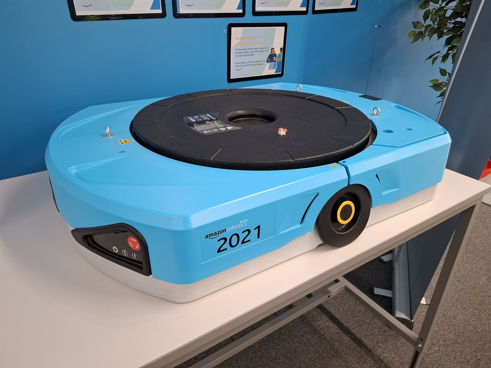 A blue Amazon Robotics drive unit, a low wheeled robot that lifts shelving pods, displayed on a table.