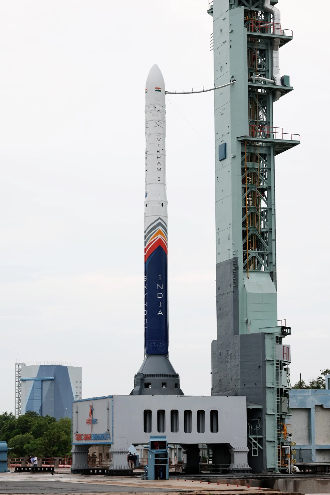 Skyroot’s Vikram-1 rocket, white with a blue and orange band, standing beside the launch tower at Sriharikota.