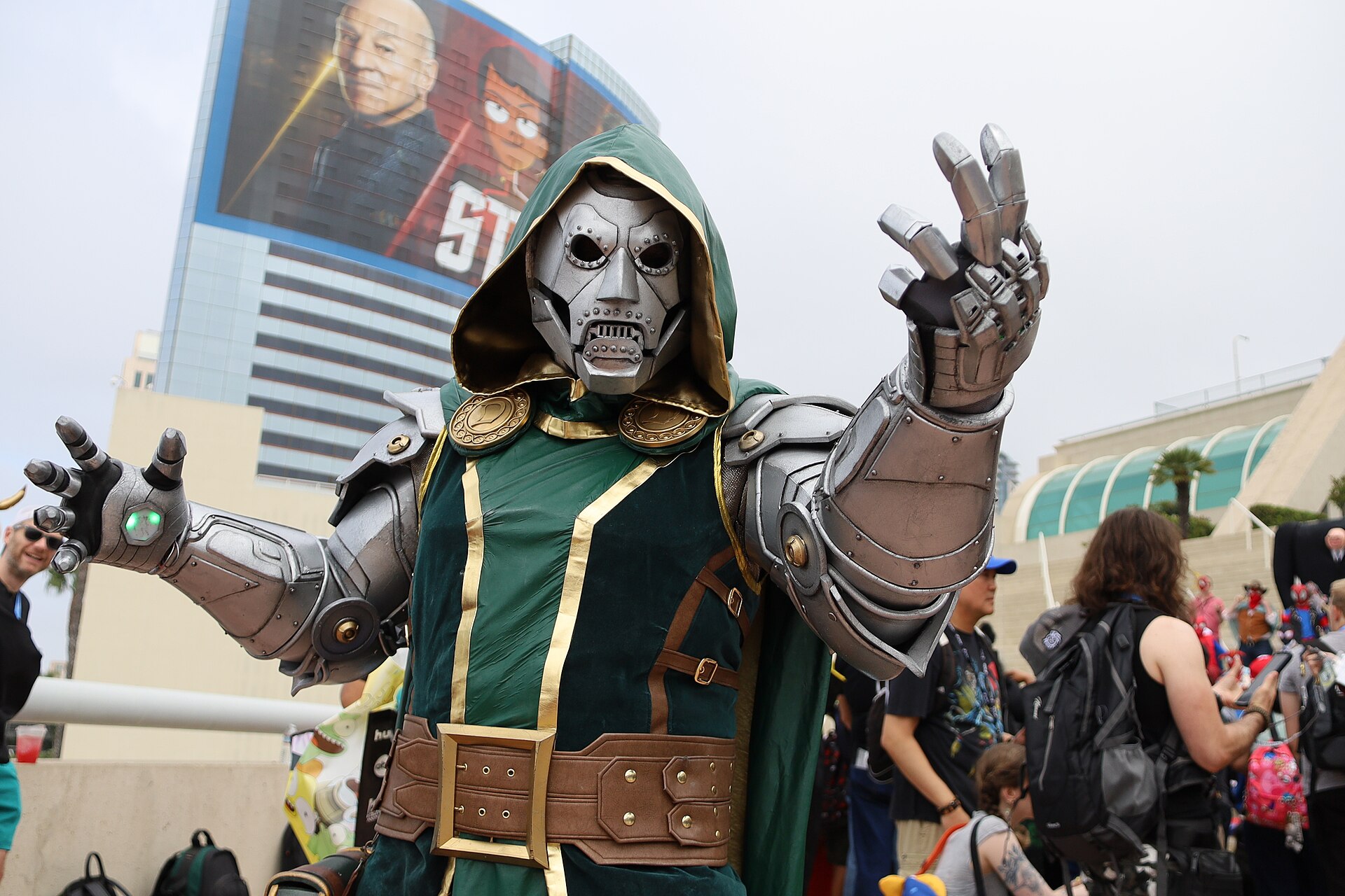 A Doctor Doom cosplayer in a detailed metal suit raises both armored hands.