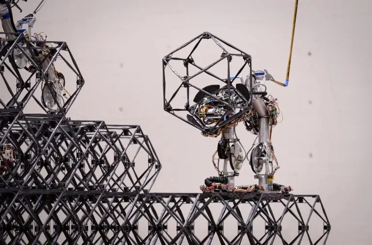 NASA’s SOLL-E robot carries an open lattice building block while standing on a structure made of matching blocks.