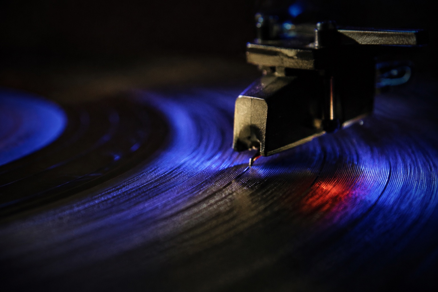 Macro photograph of a record-player stylus resting in a vinyl groove under cobalt and red light.