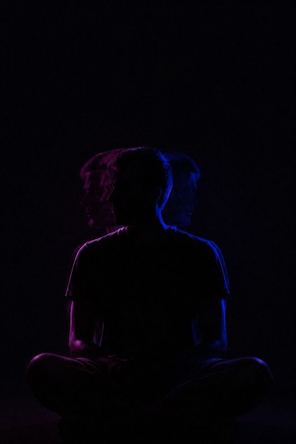 A dark long-exposure silhouette of a seated person with faint magenta and blue head traces.