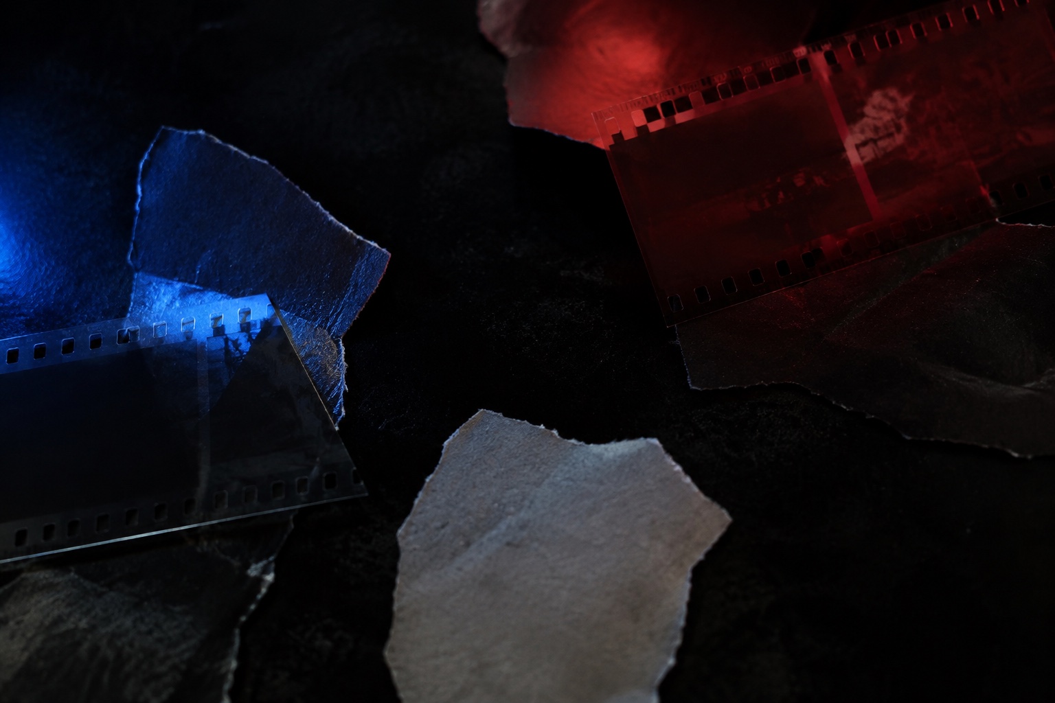 Translucent photographic negatives and torn blank paper overlapping on a black surface under blue and red light.