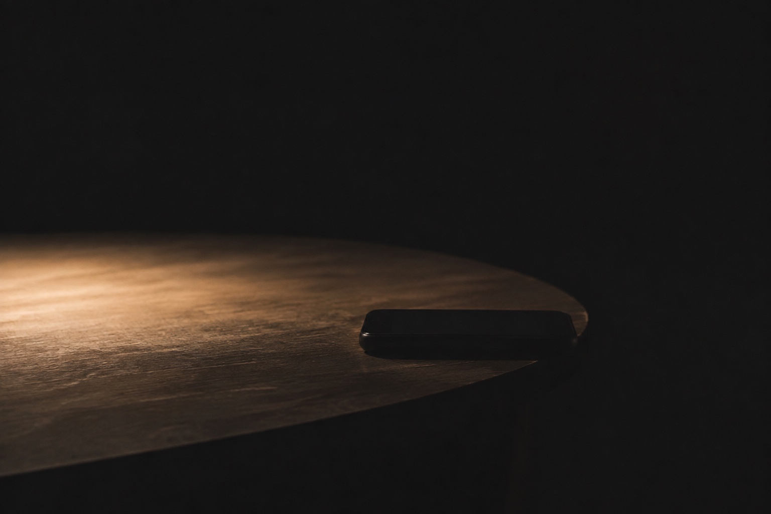 A plain smartphone lying face down at the edge of a dark table, outside a small pool of warm light.