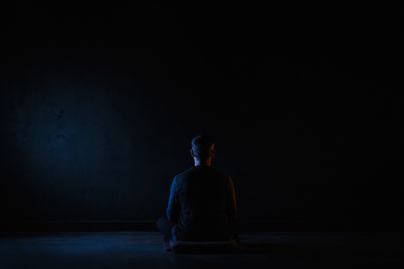 An adult seen from behind, sitting on a cushion in a nearly black room with faint blue light.