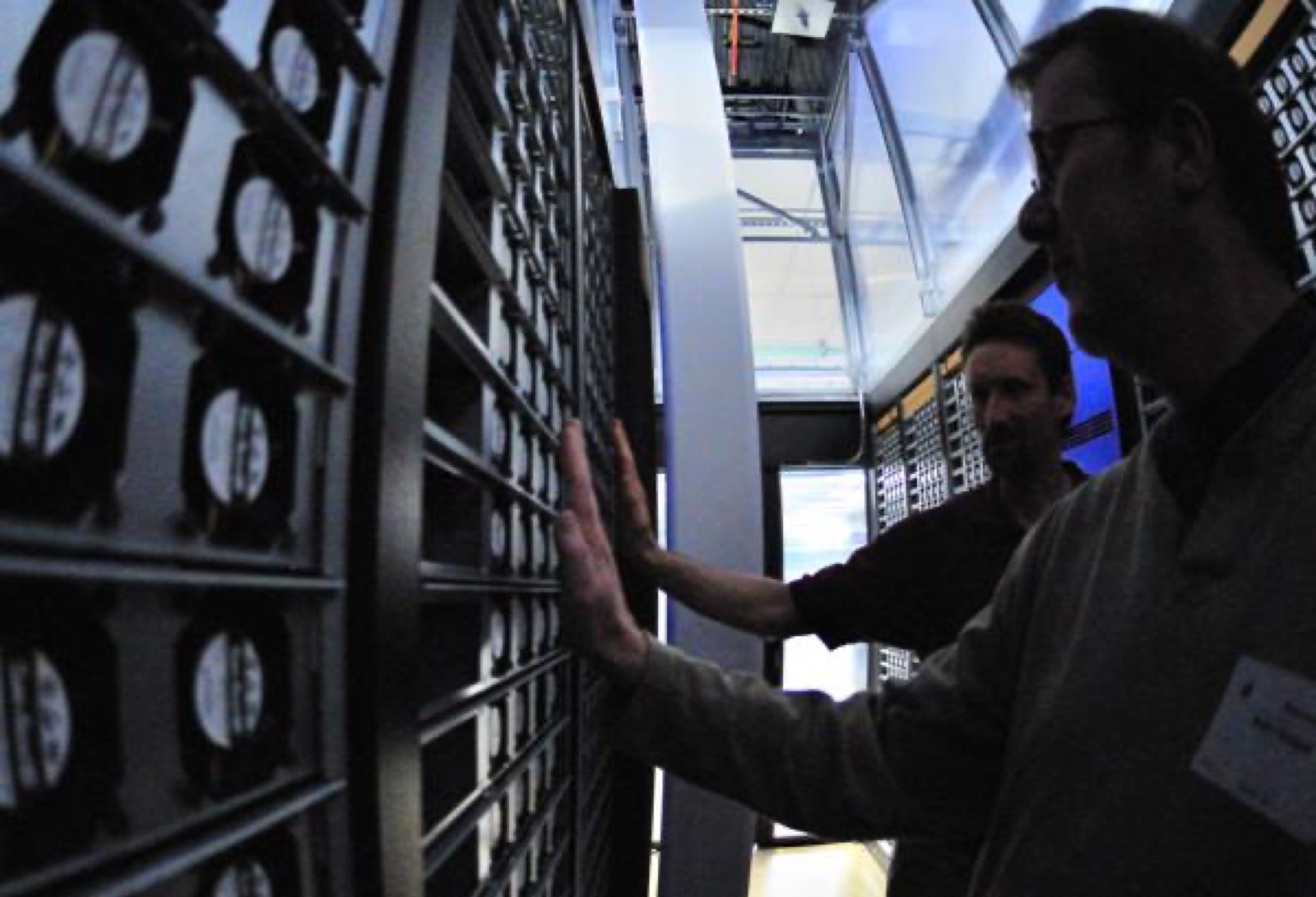 Two engineers in the hot aisle of a data center, hand resting on a rack of servers, cooling fans and cabling receding into the dark