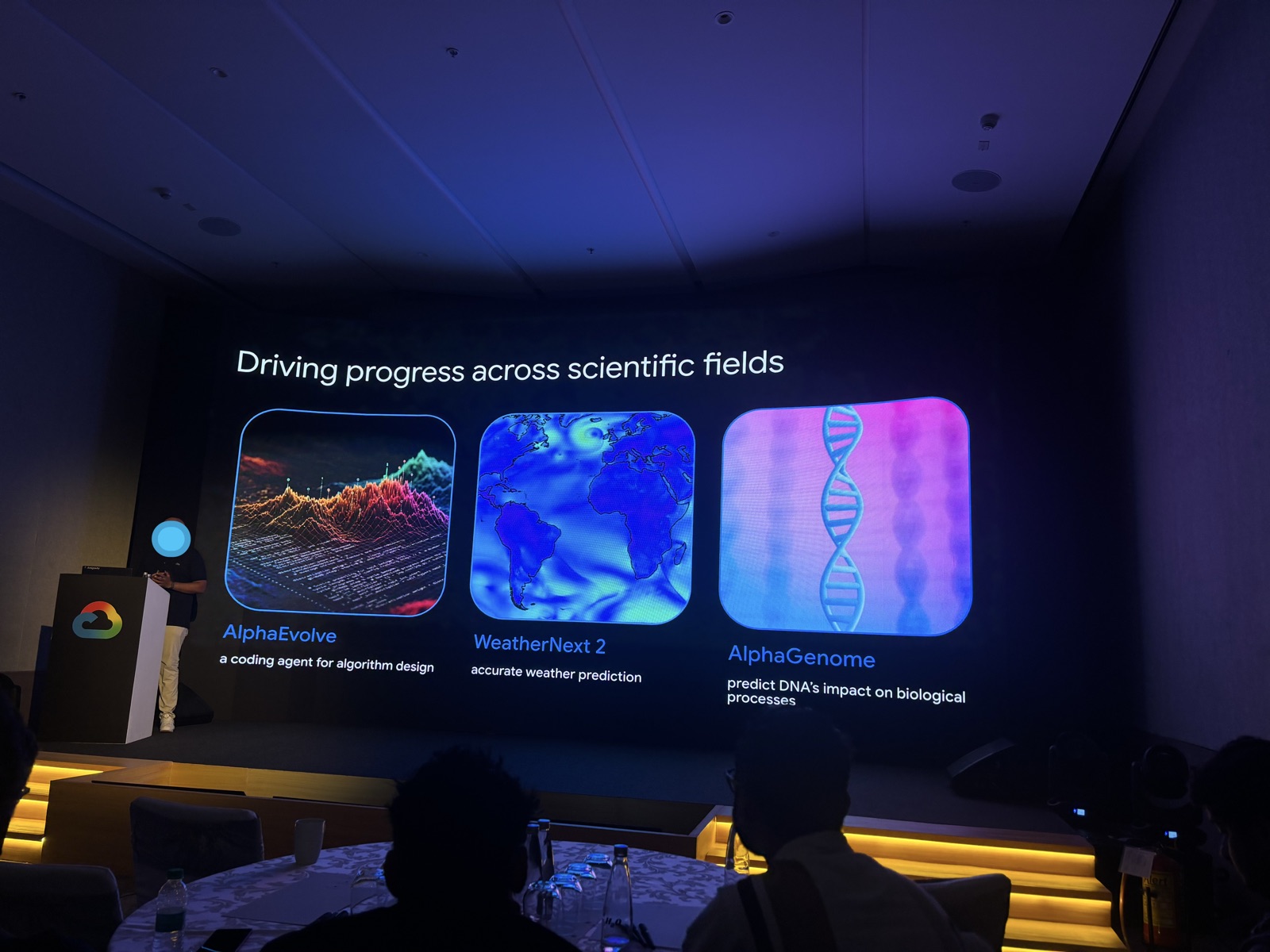The stage at Google's CTO Connect: a speaker at a Google Cloud podium beside a large screen reading Driving progress across scientific fields, showing AlphaEvolve, WeatherNext 2, and AlphaGenome, above the silhouettes of a seated audience.