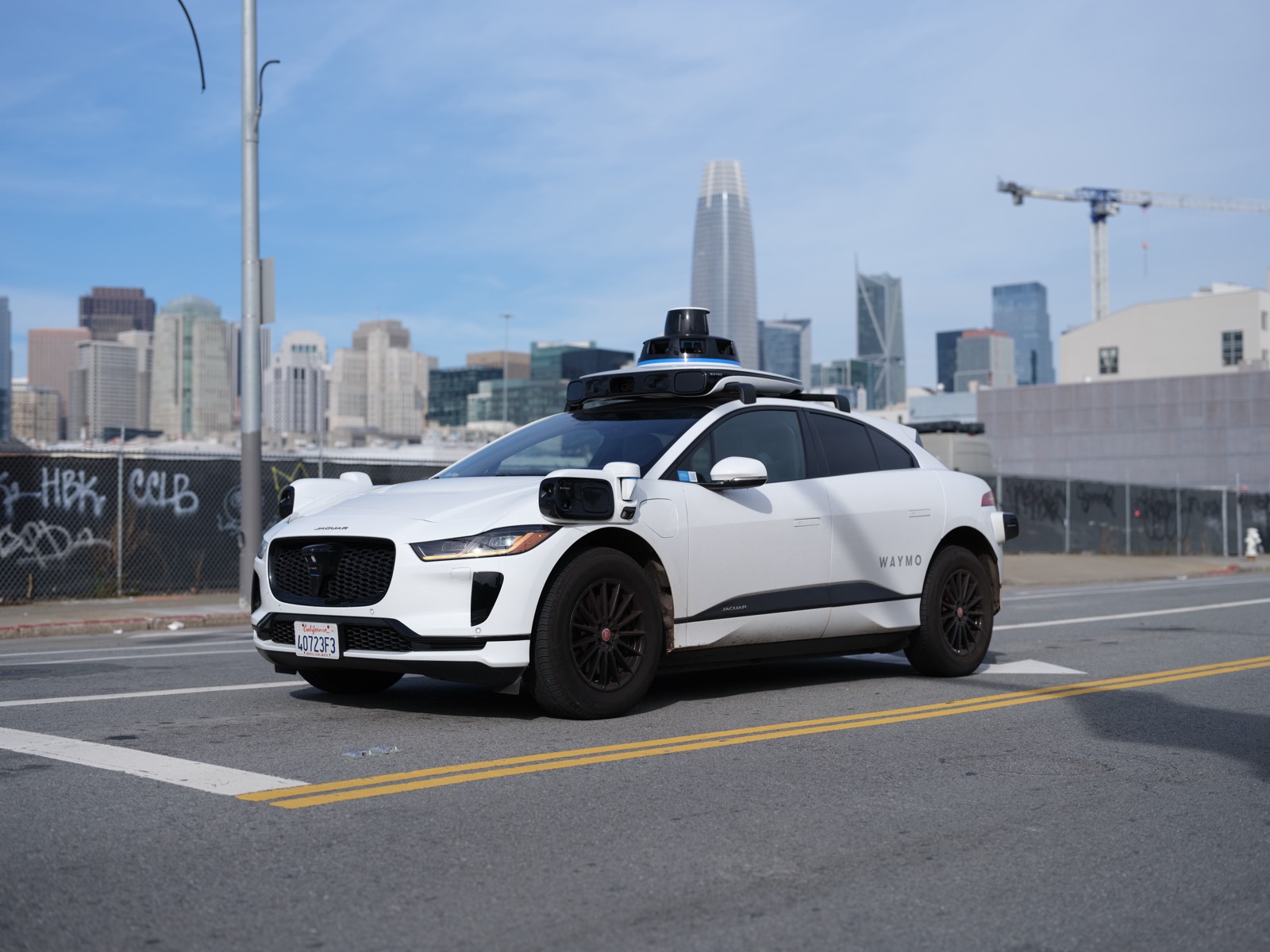A white Waymo autonomous Jaguar with a roof-mounted sensor array driving on a city street.
