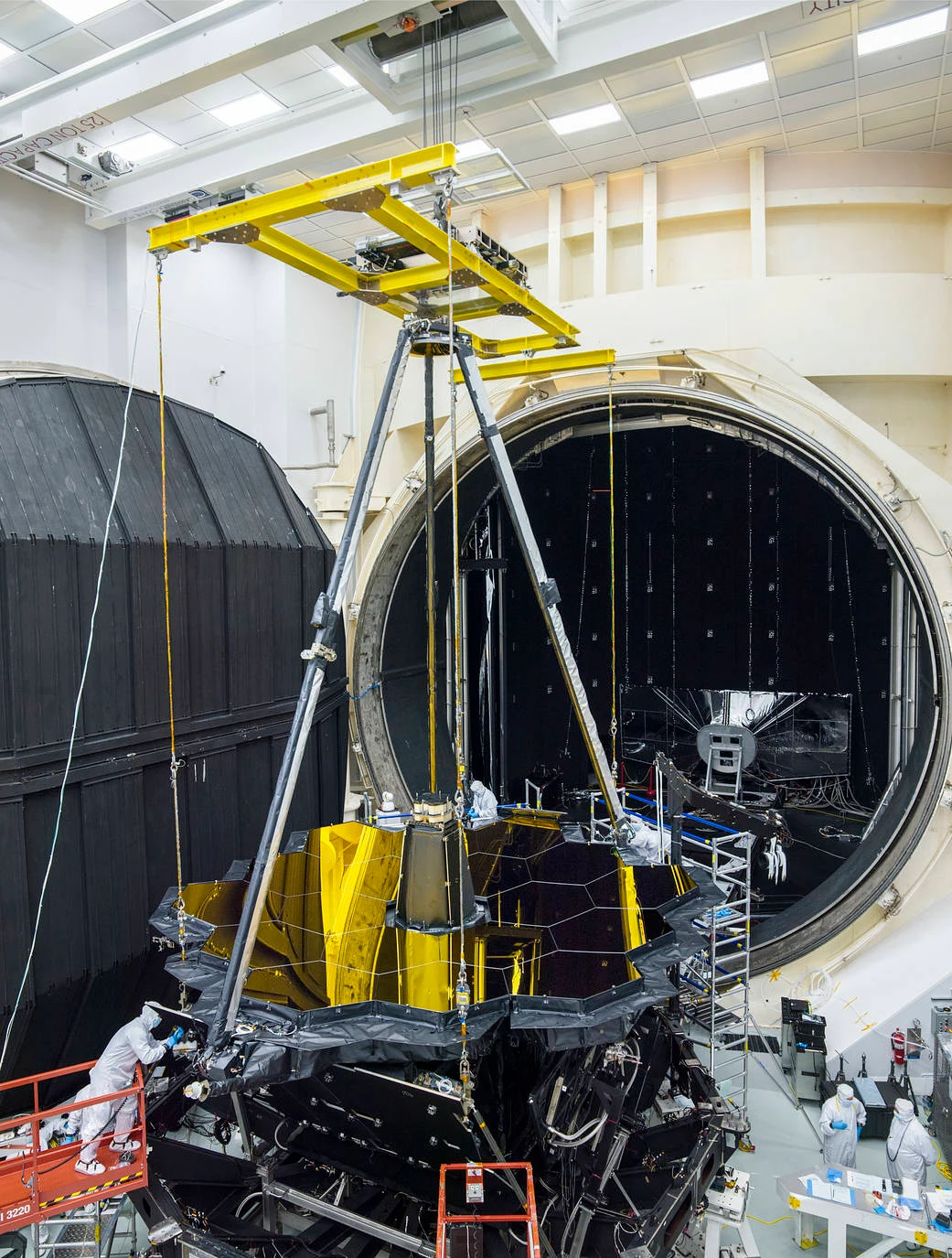 The James Webb Space Telescope suspended in front of the large circular opening of NASA’s Chamber A, with technicians below